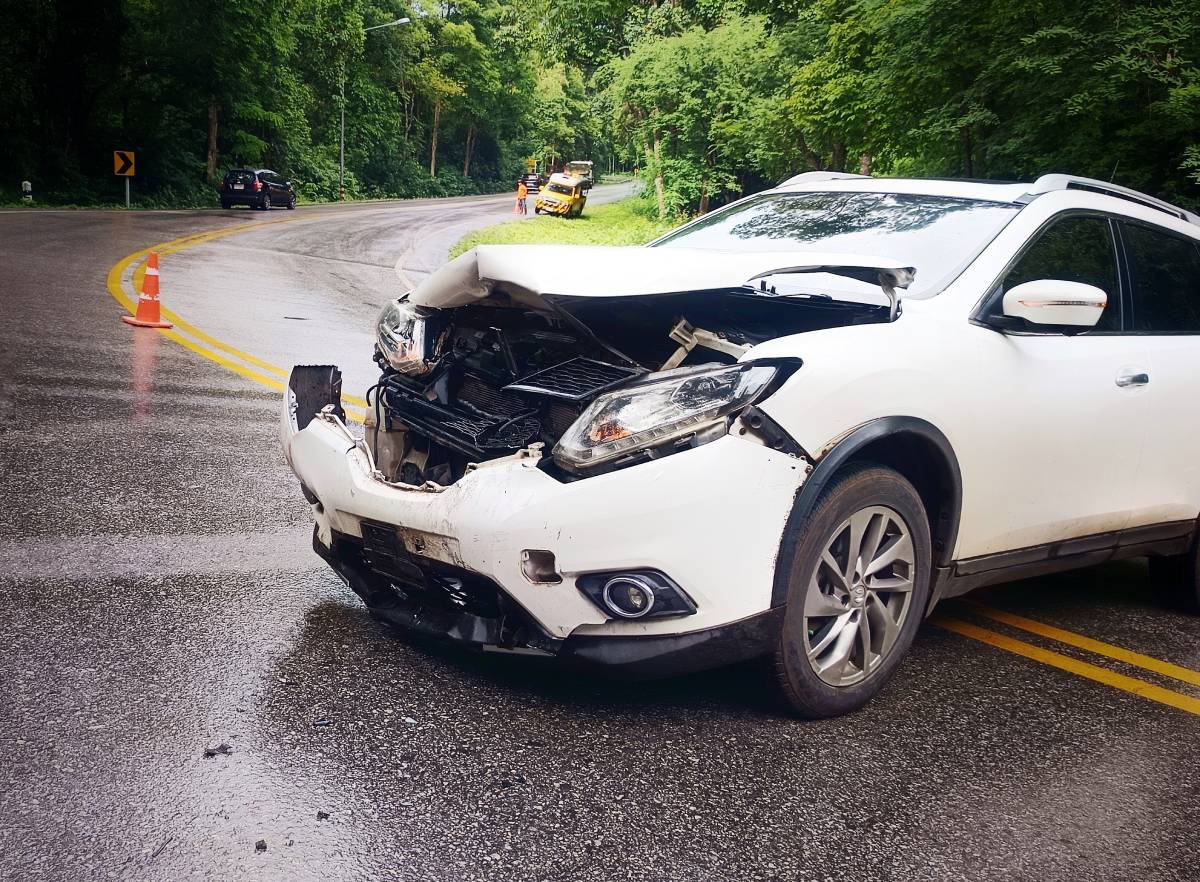 a white car totaled in an accident on a rainy road surrounded by trees